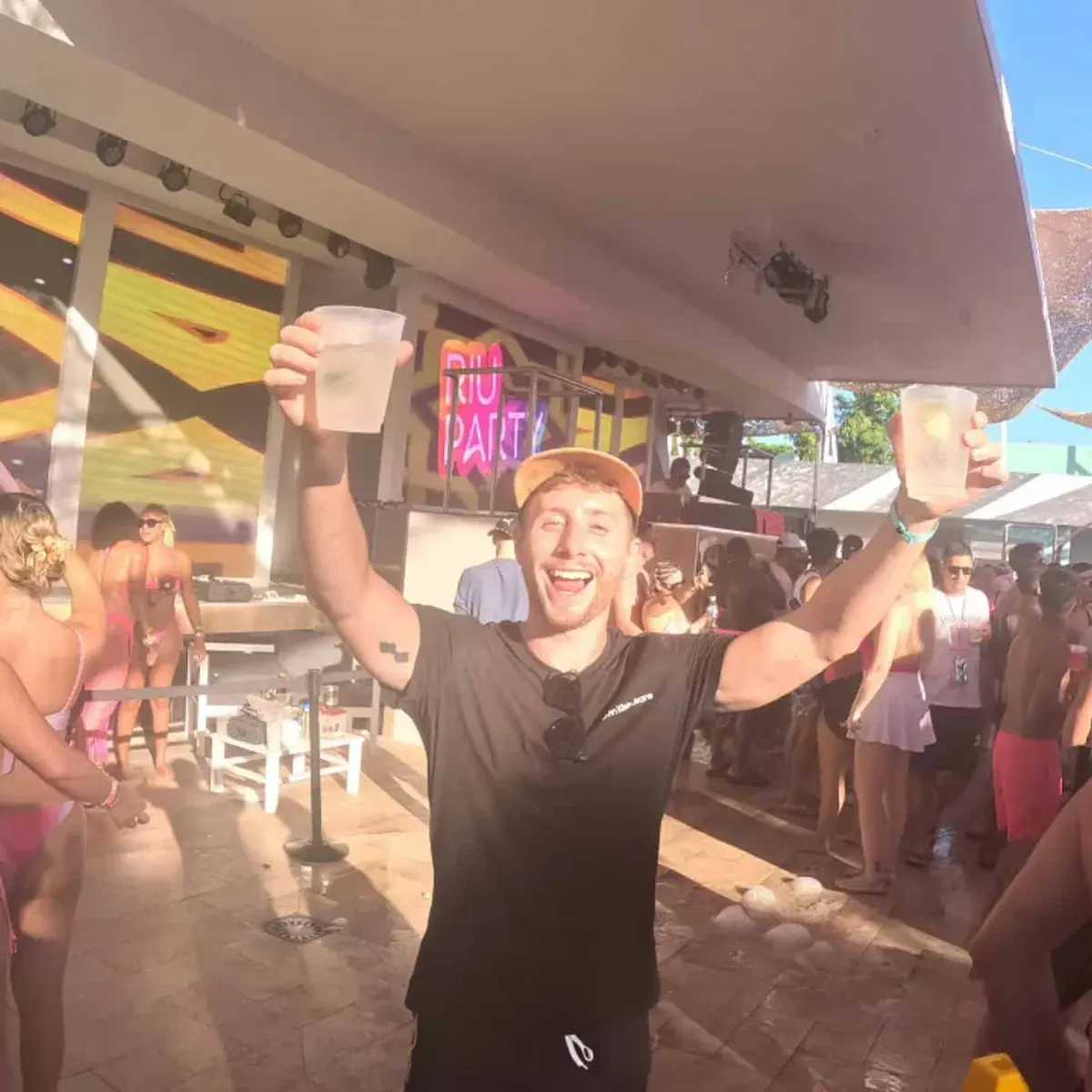 Vibrant pool party scene with cheerful man holding drinks in the air, lively crowd, and colorful lighting.