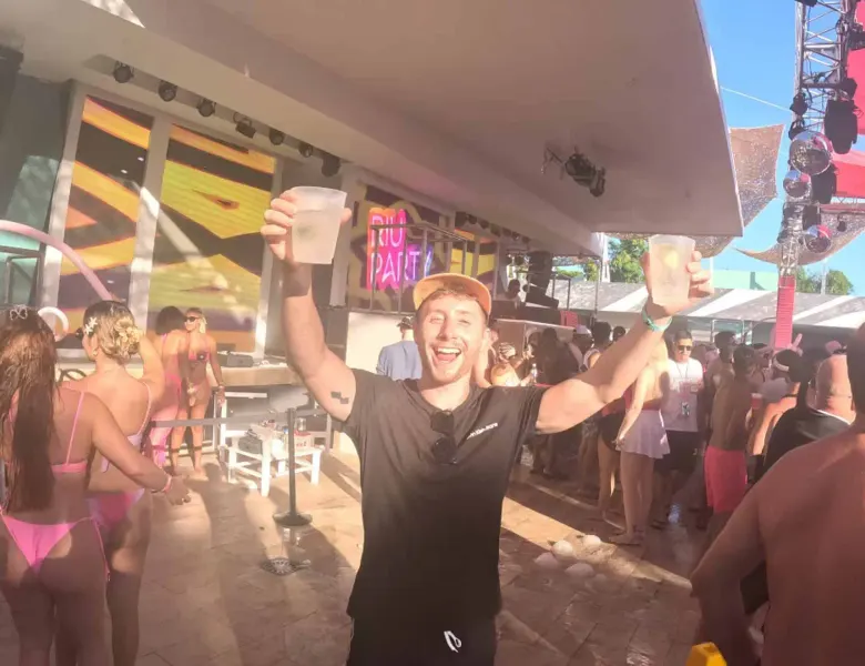 Vibrant pool party scene with cheerful man holding drinks in the air, lively crowd, and colorful lighting.