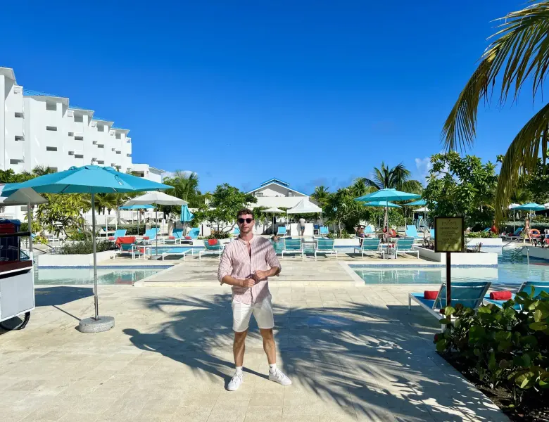 Man standing by pool with umbrellas, lounge chairs, and palm trees