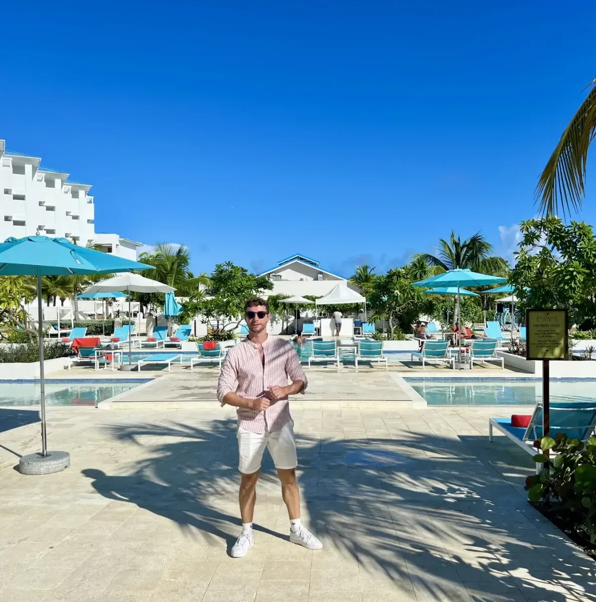 Man standing by pool with umbrellas, lounge chairs, and palm trees