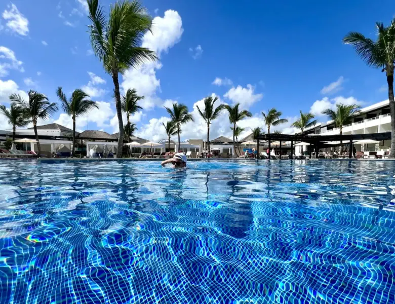 Person in pool, palm trees, resort buildings, blue sky