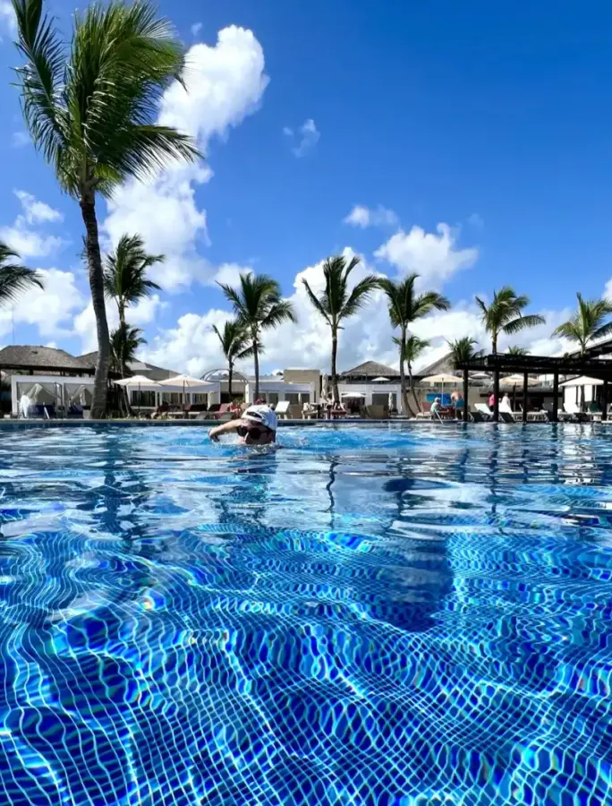 Person in pool, palm trees, resort buildings, blue sky