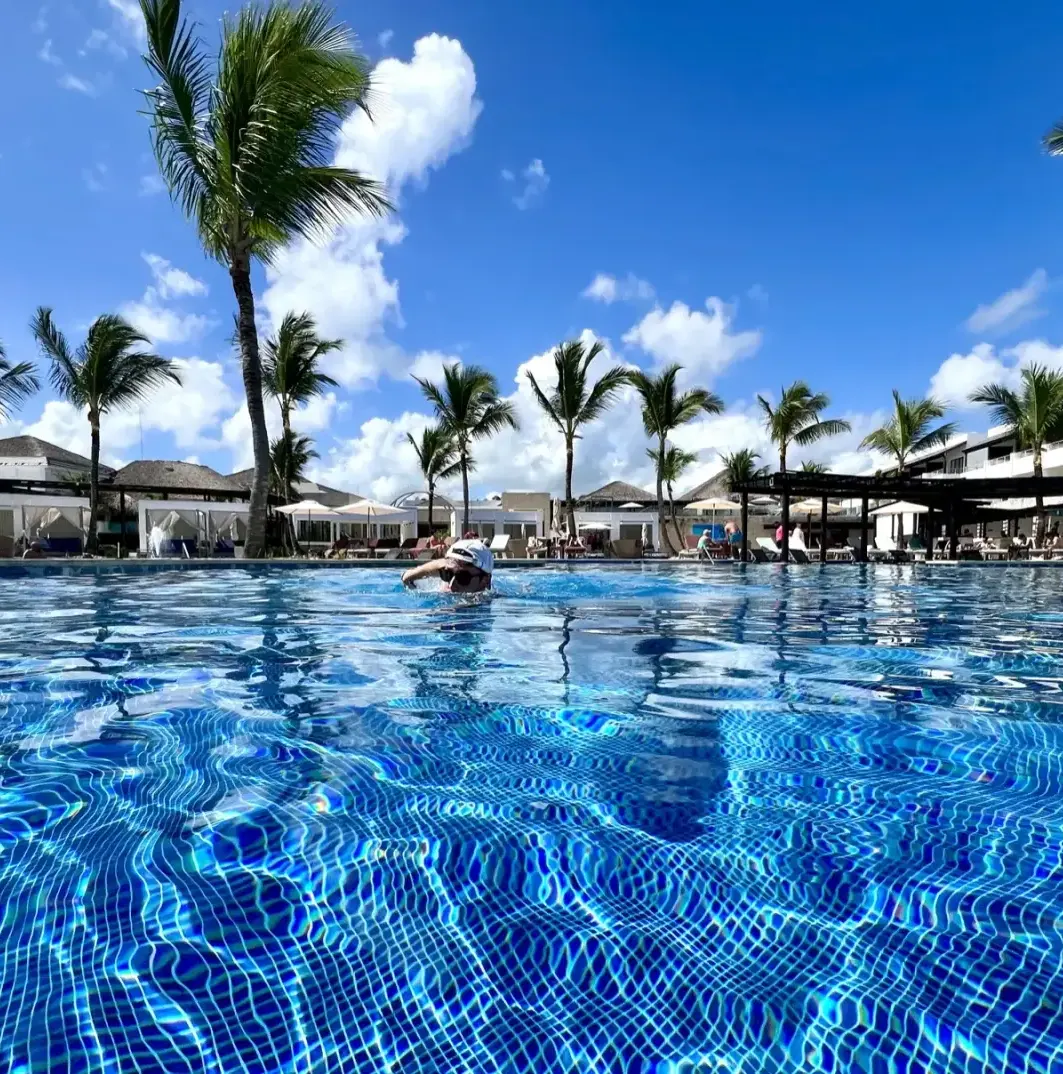 Person in pool, palm trees, resort buildings, blue sky