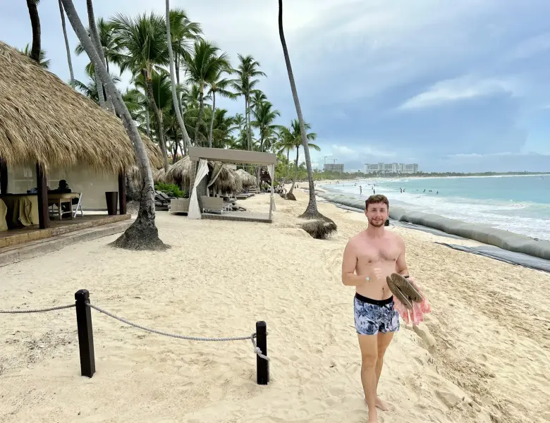 Man on sandy beach with palm trees, thatched structures, and ocean