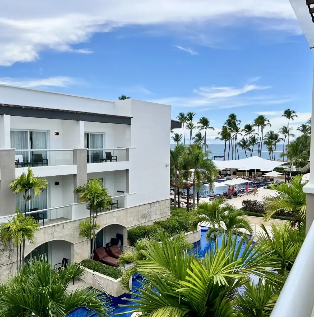 Balcony view of resort buildings, pool, palm trees, and beach