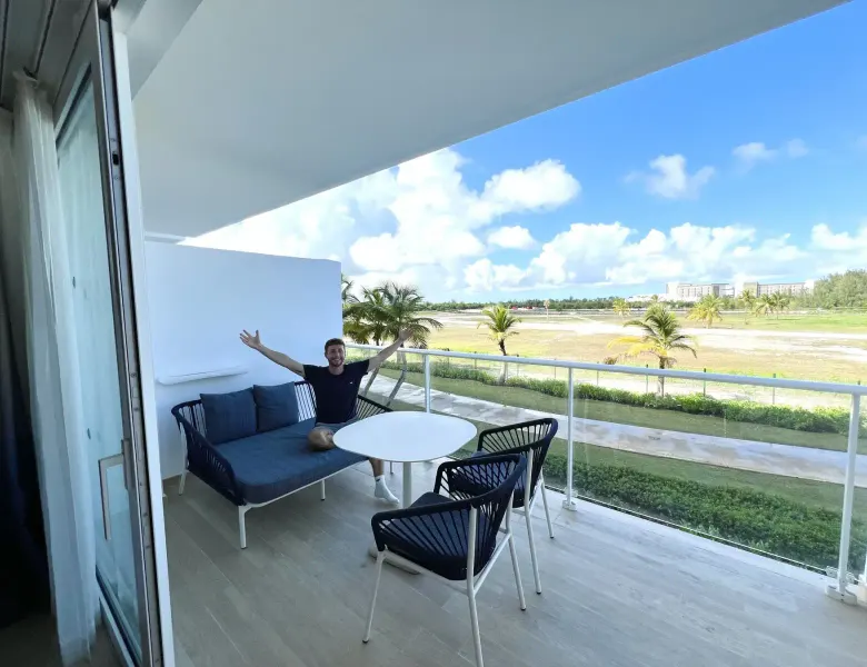 Man on balcony with sofa, table, chairs, palm trees, grass, building