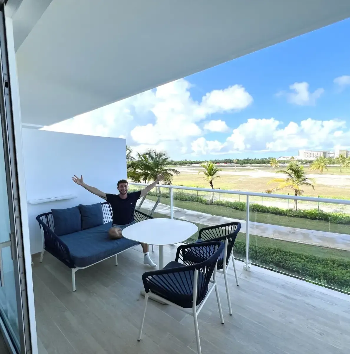 Man on balcony with sofa, table, chairs, palm trees, grass, building