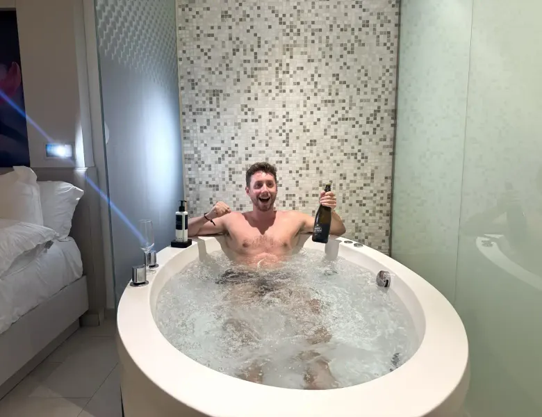 Man in jacuzzi with bottle, bed and tiled wall in background