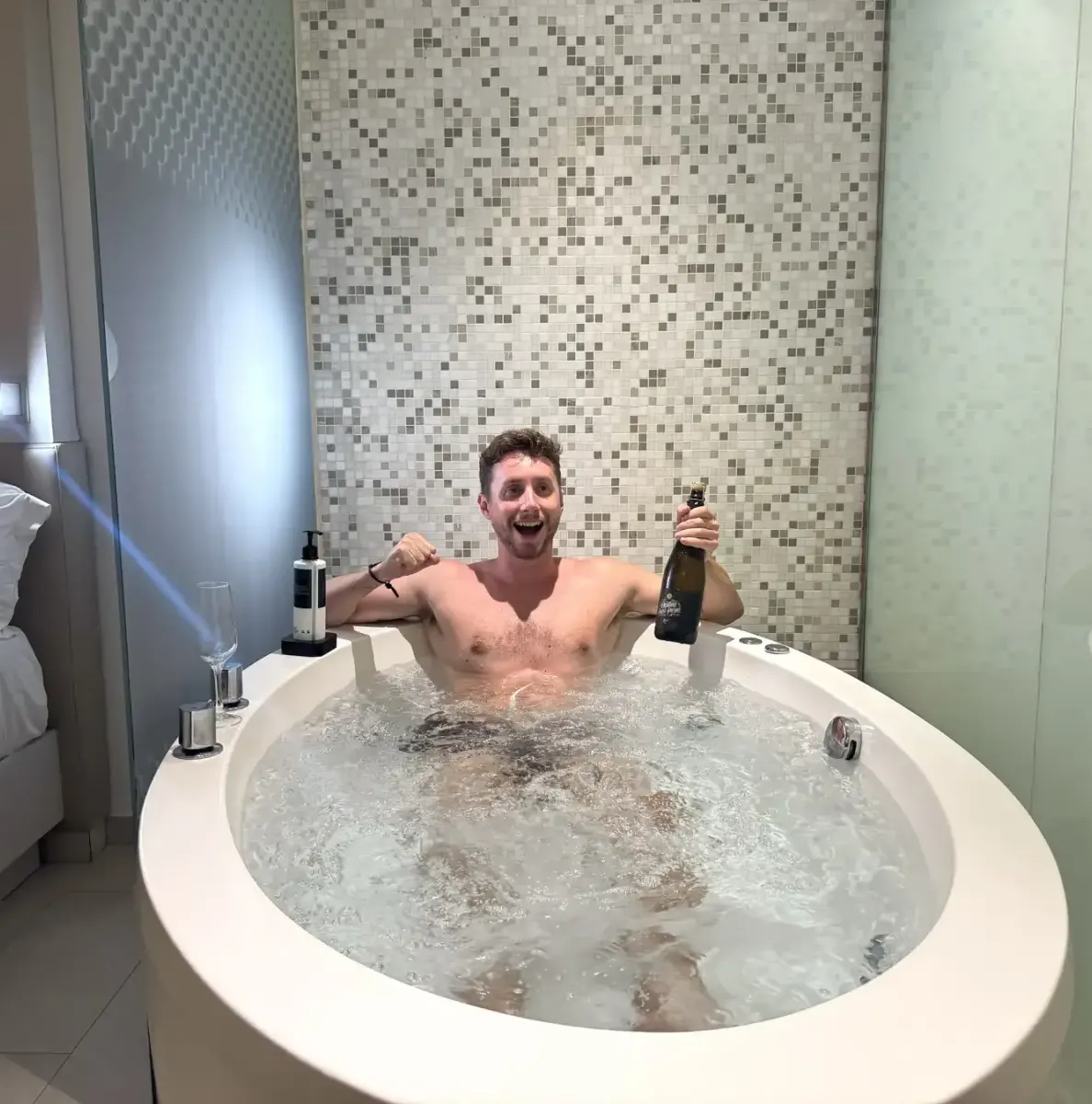 Man in jacuzzi with bottle, bed and tiled wall in background