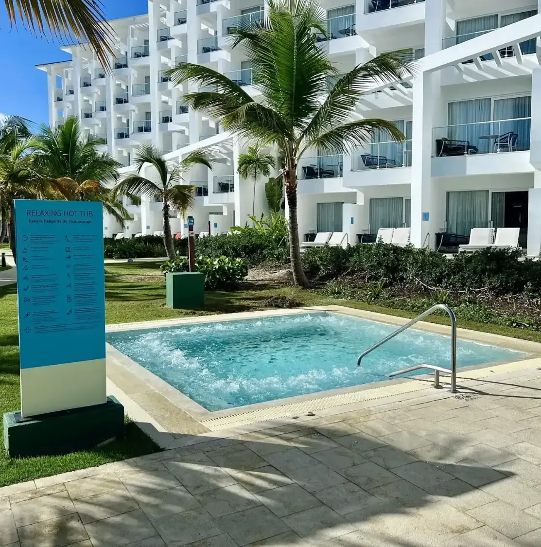 Jacuzzi with sign, palm trees, hotel building, lounge chairs, paved area