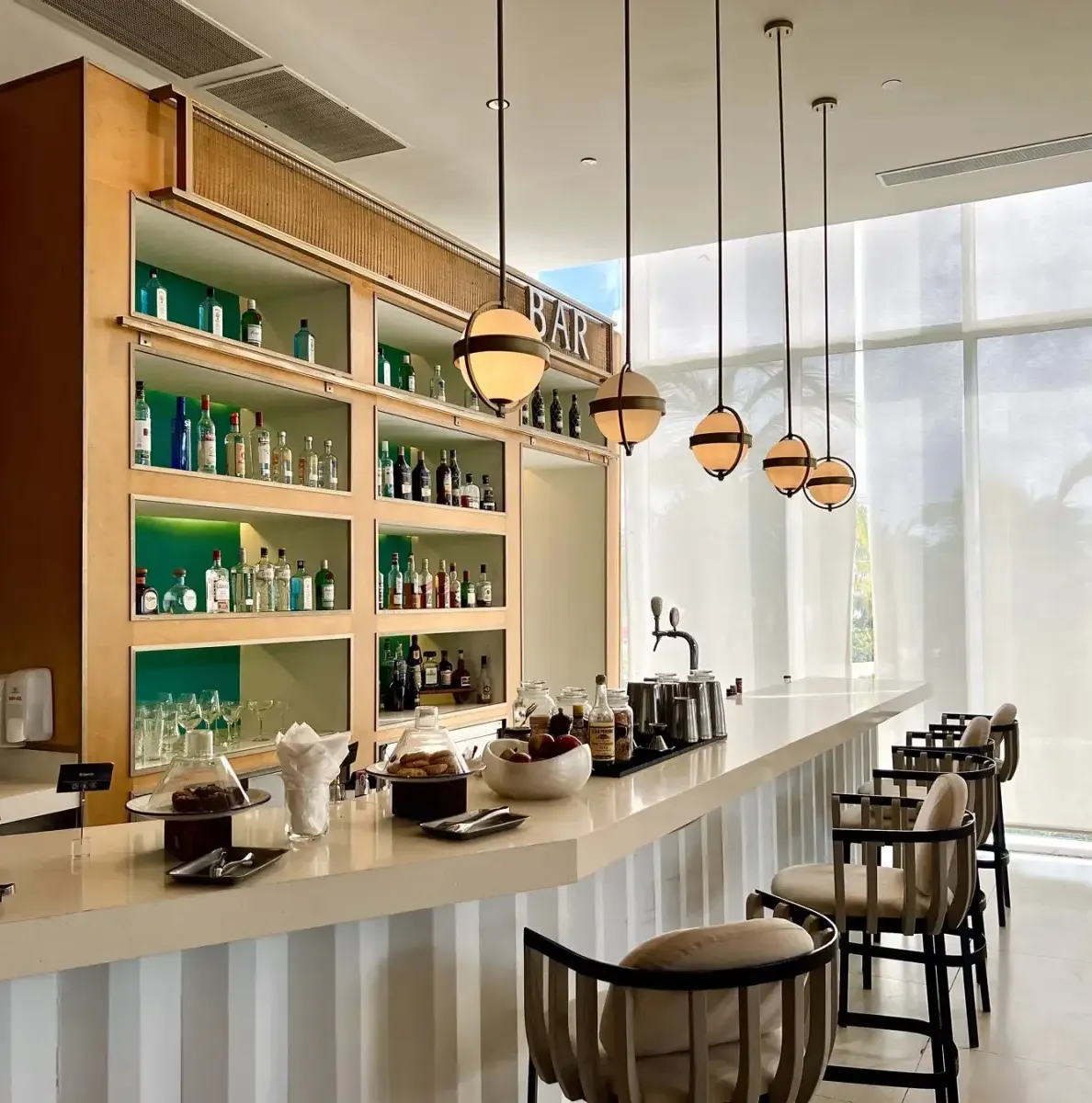 bar counter with stools, shelves of bottles, hanging lights, large windows