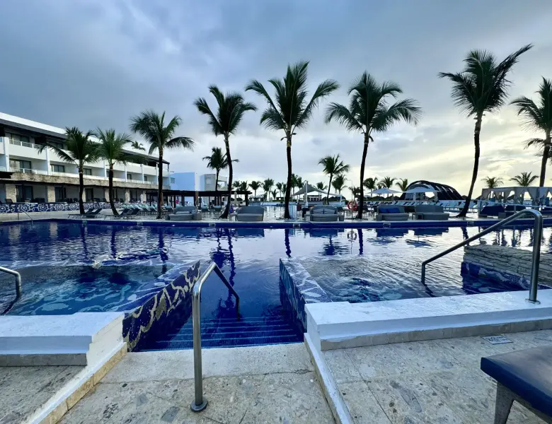 Pool with steps, palm trees, and resort building in background