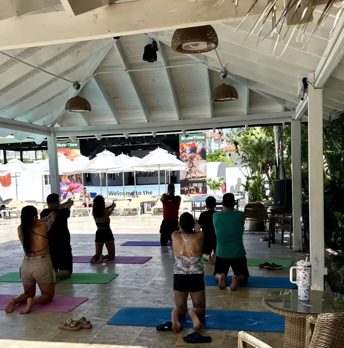 People doing yoga under a covered area near a pool with umbrellas
