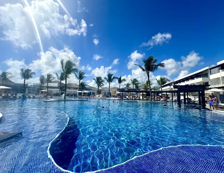Large pool with blue water, palm trees, resort buildings, people swimming