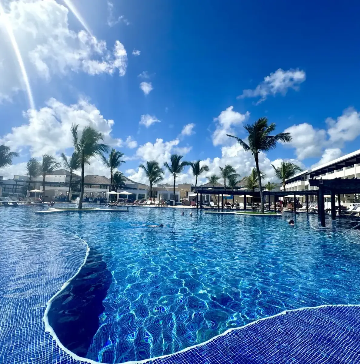 Large pool with blue water, palm trees, resort buildings, people swimming
